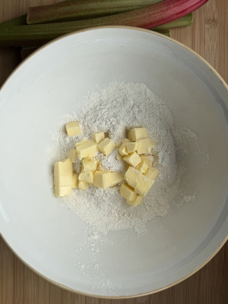 A large white bowl containing flour and cubed butter, ready for mixing, sits on a wooden surface with stalks of rhubarb in the background.