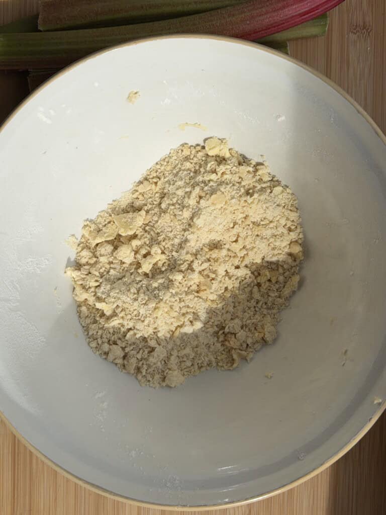A white bowl contains crumbly flour and butter mixture, likely for making pastry or crumble, on a wooden surface with rhubarb stalks partially visible at the top.