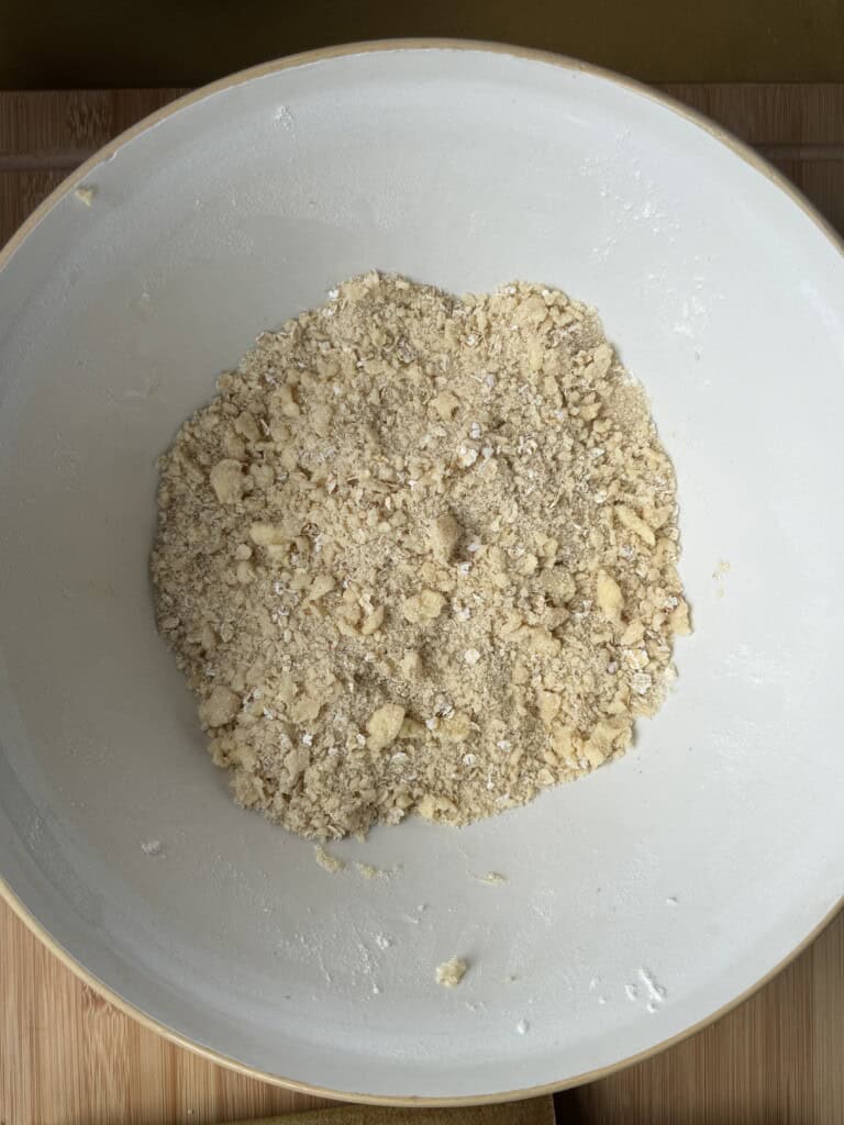A white mixing bowl containing a crumbly mixture of flour, oats, and butter sits on a wooden surface, likely for making a crumble topping or pastry dough.