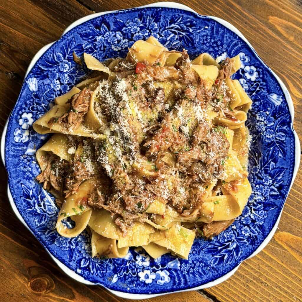 Wide pappardelle pasta with a rich lamb ragù and grated cheese is served on a blue and white floral plate atop a wooden table.