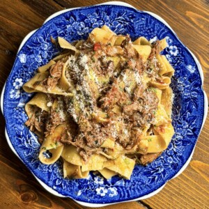 Wide pappardelle pasta with a rich lamb ragù and grated cheese is served on a blue and white floral plate atop a wooden table.