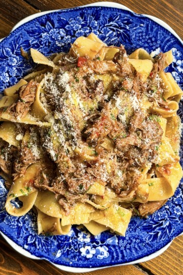 Wide pappardelle pasta with a rich lamb ragù and grated cheese is served on a blue and white floral plate atop a wooden table.