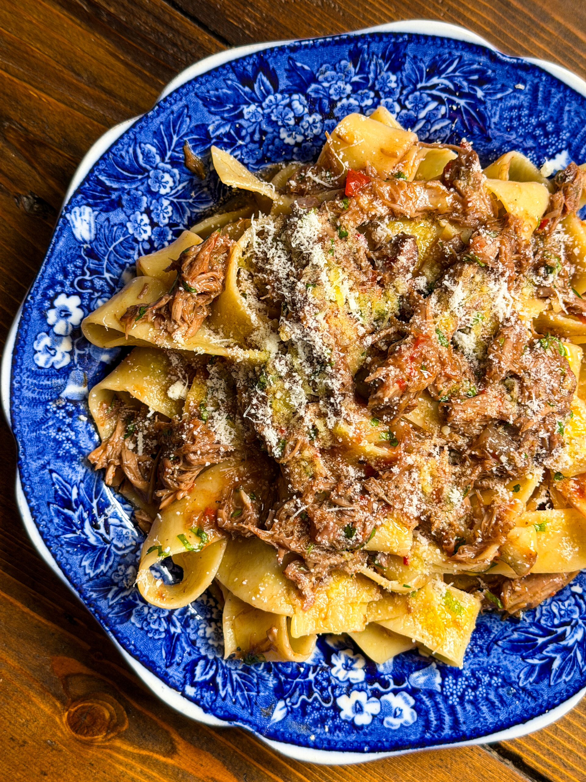 Wide pasta noodles topped with rich lamb ragù and sprinkled with grated parmesan, served on a blue floral-patterned plate on a wooden table.