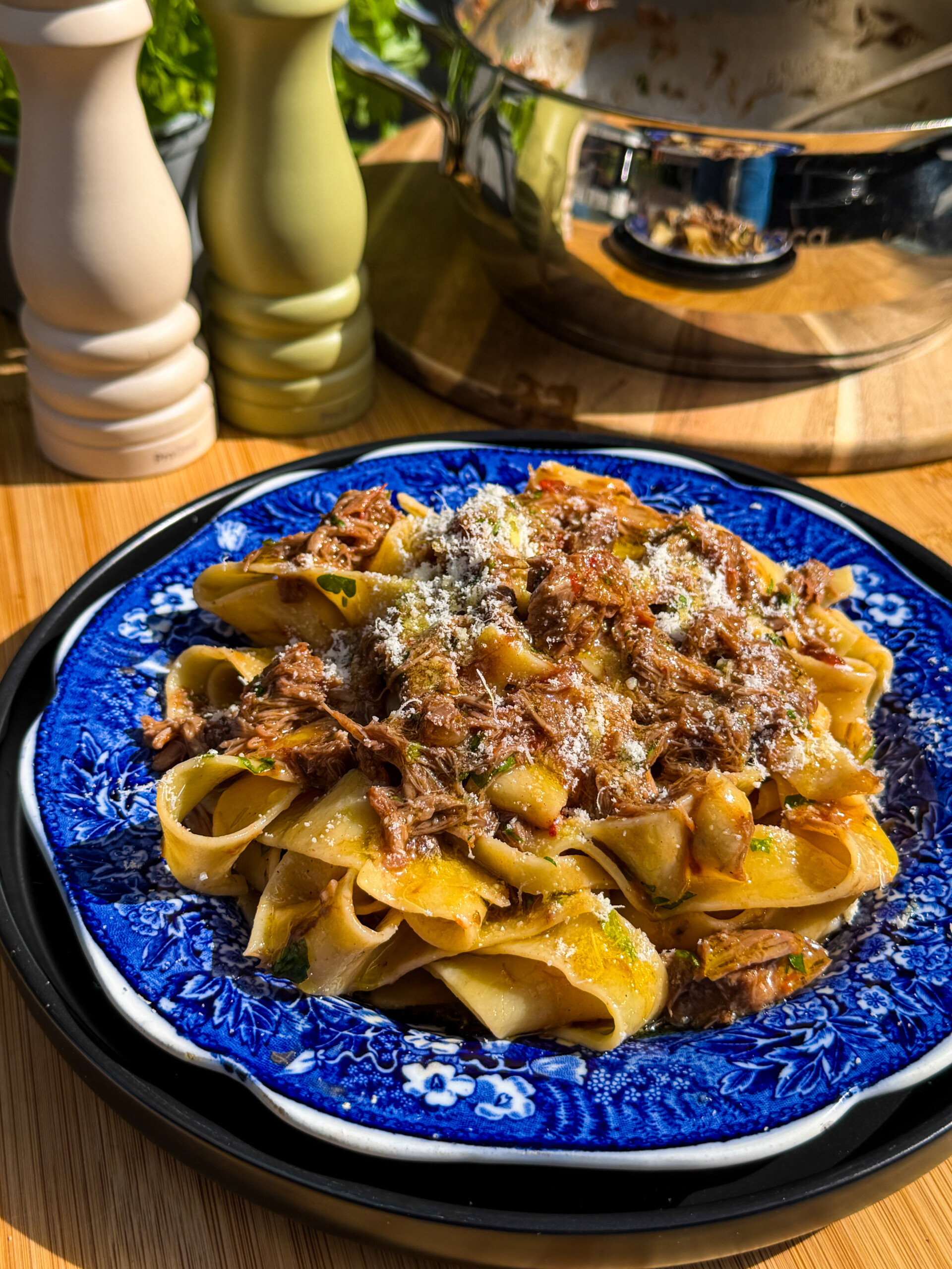 Wide pappardelle pasta topped with rich lamb ragù and grated cheese on a blue plate. Sunlight highlights the dish. A saucepan, salt and pepper shakers, and a wooden surface are in the background.