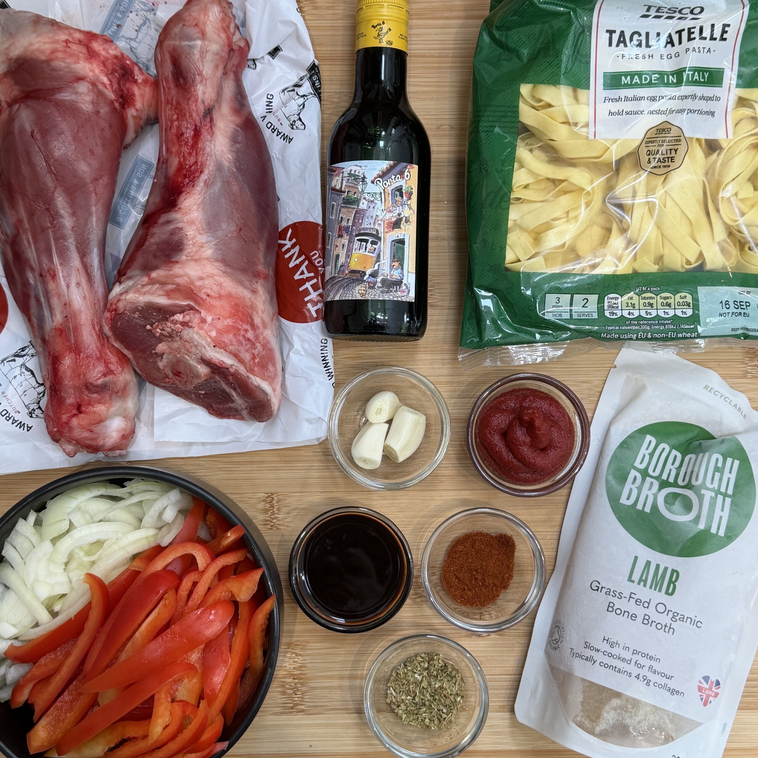 Ingredients for cooking a rich lamb ragù: raw lamb shanks, a bottle of red wine, dried tagliatelle pasta, sliced onion, red pepper, garlic cloves, tomato purée, spices, herb mix, a bowl of stock, and a pack of lamb bone stock arranged on a wooden surface.