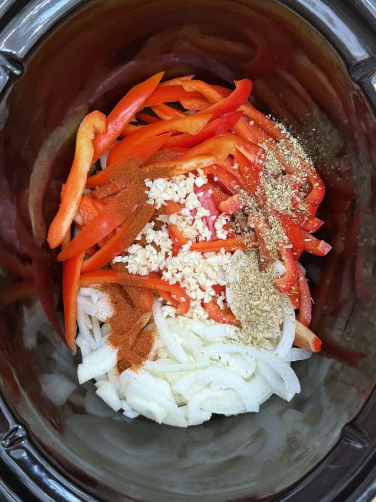 Thinly sliced red peppers, onions, minced garlic, paprika, and dried herbs are layered inside a black slow cooker.