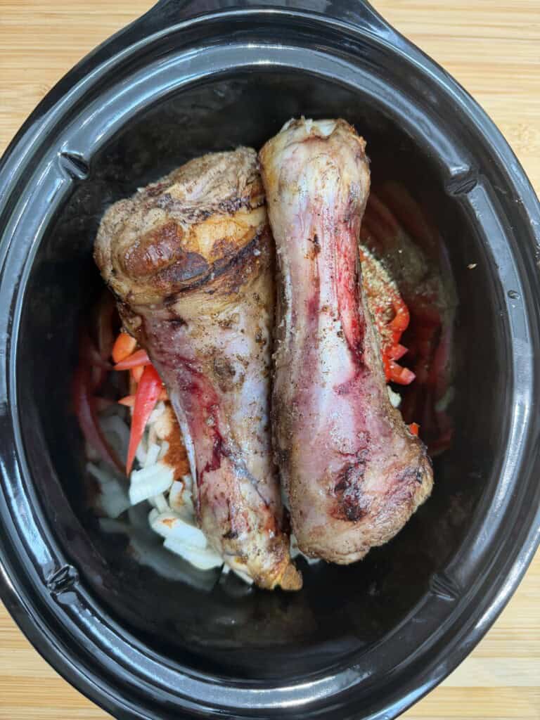Two large cooked bones with meat rest on chopped onions and red peppers inside a black slow cooker, ready for further cooking.