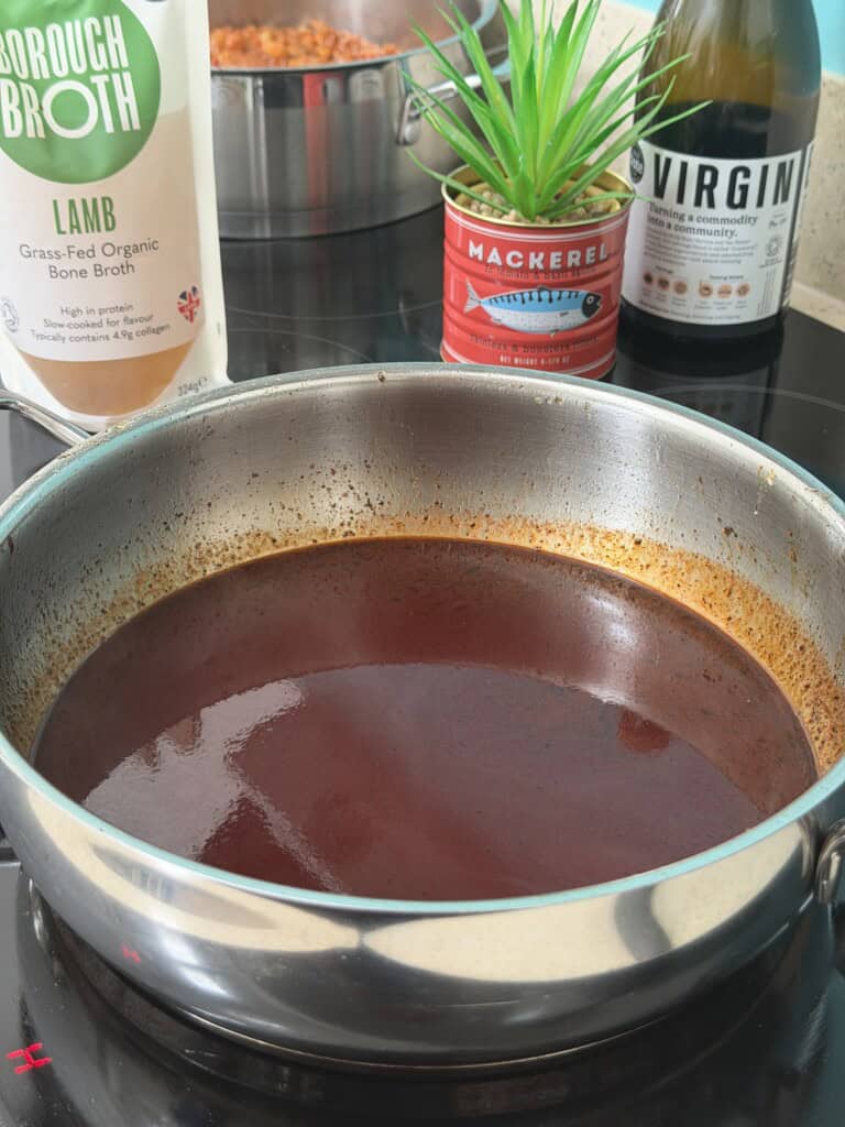 A stainless steel pan with dark brown sauce on a hob, with a carton of bone broth, a plant in a mackerel tin, and a bottle of vinegar in the background.