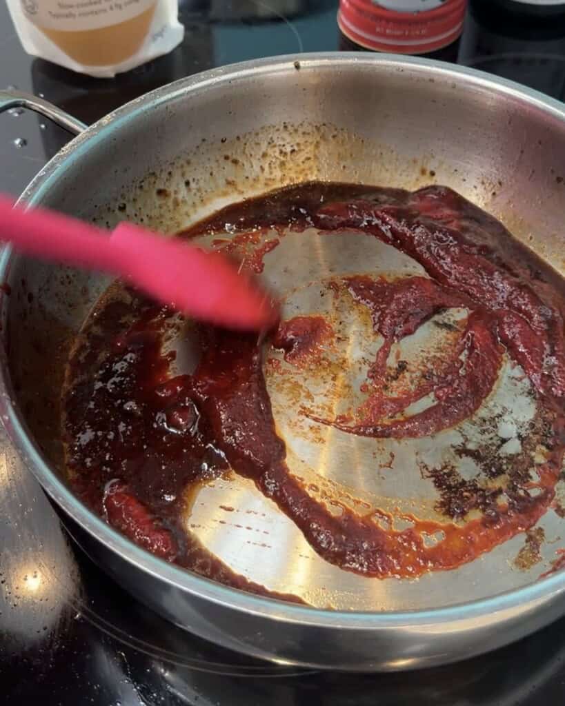A pink spatula stirs a deep red sauce, possibly tomato purée, in a stainless steel pan on a cooker. The sauce is partially mixed, with some browning and caramelisation visible on the bottom of the pan.