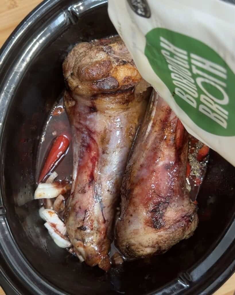 Two large beef bones, onions, and red chilli peppers in a slow cooker, with someone pouring bone broth from a labelled container over the ingredients.