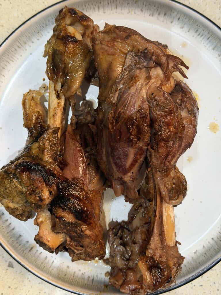 Two pieces of cooked lamb shank with browned, crispy skin and exposed bone are served on a white plate with a thin dark rim. The meat looks tender and juicy.