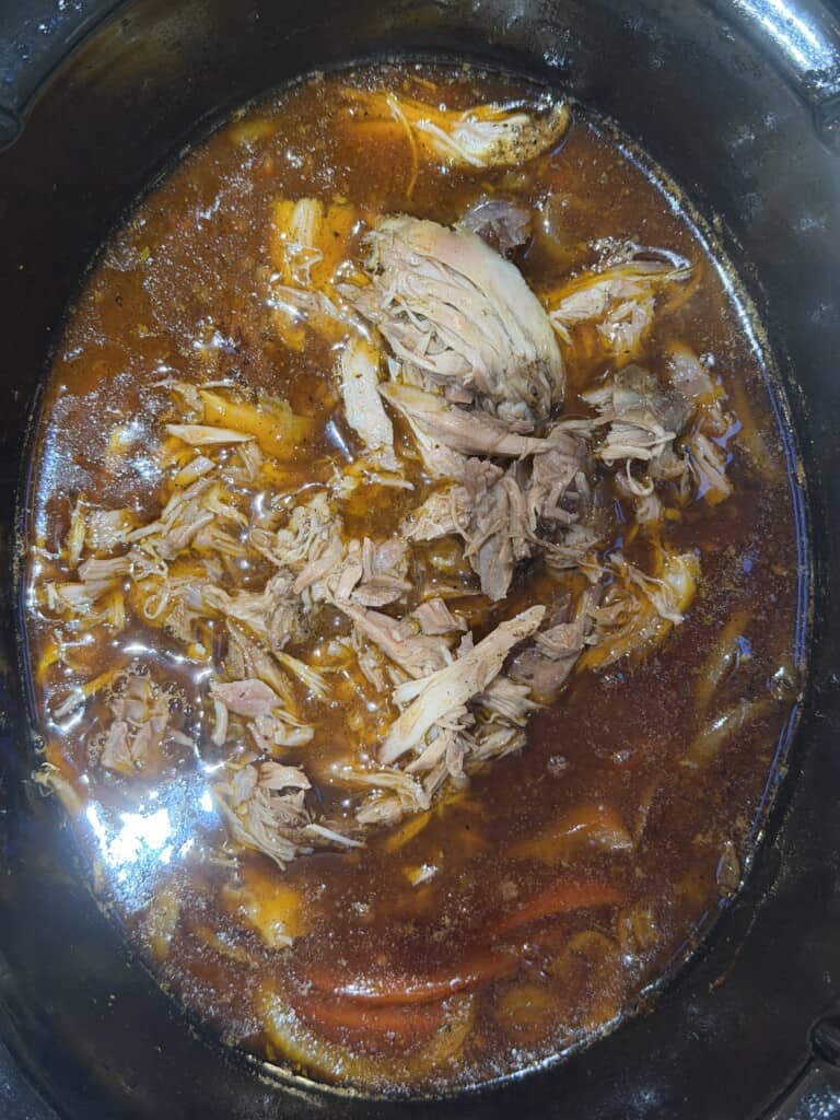 Shredded lamb in a rich, brown broth with visible spices and pieces of onion and red pepper, cooking in a black slow cooker—reminiscent of a classic ragù with its hearty flavours and aromatic vegetables.