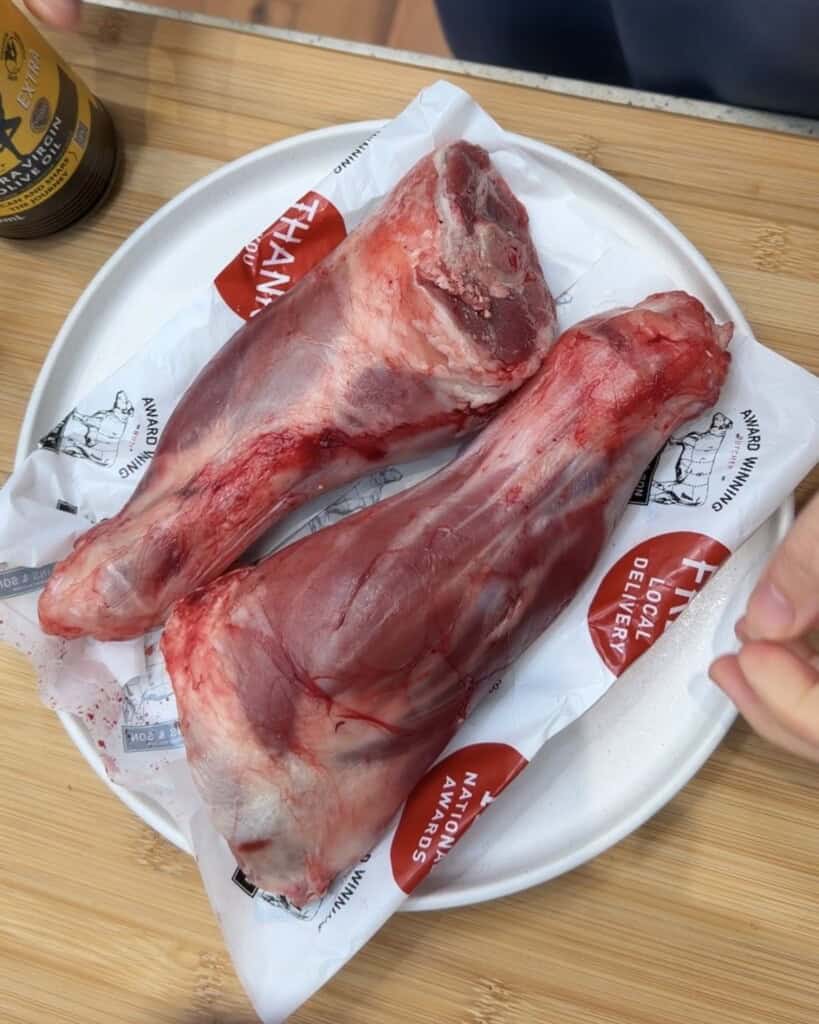 Two raw lamb shanks on butcher’s paper rest on a white plate atop a wooden surface, with a partial view of a hand and a drink in the background.