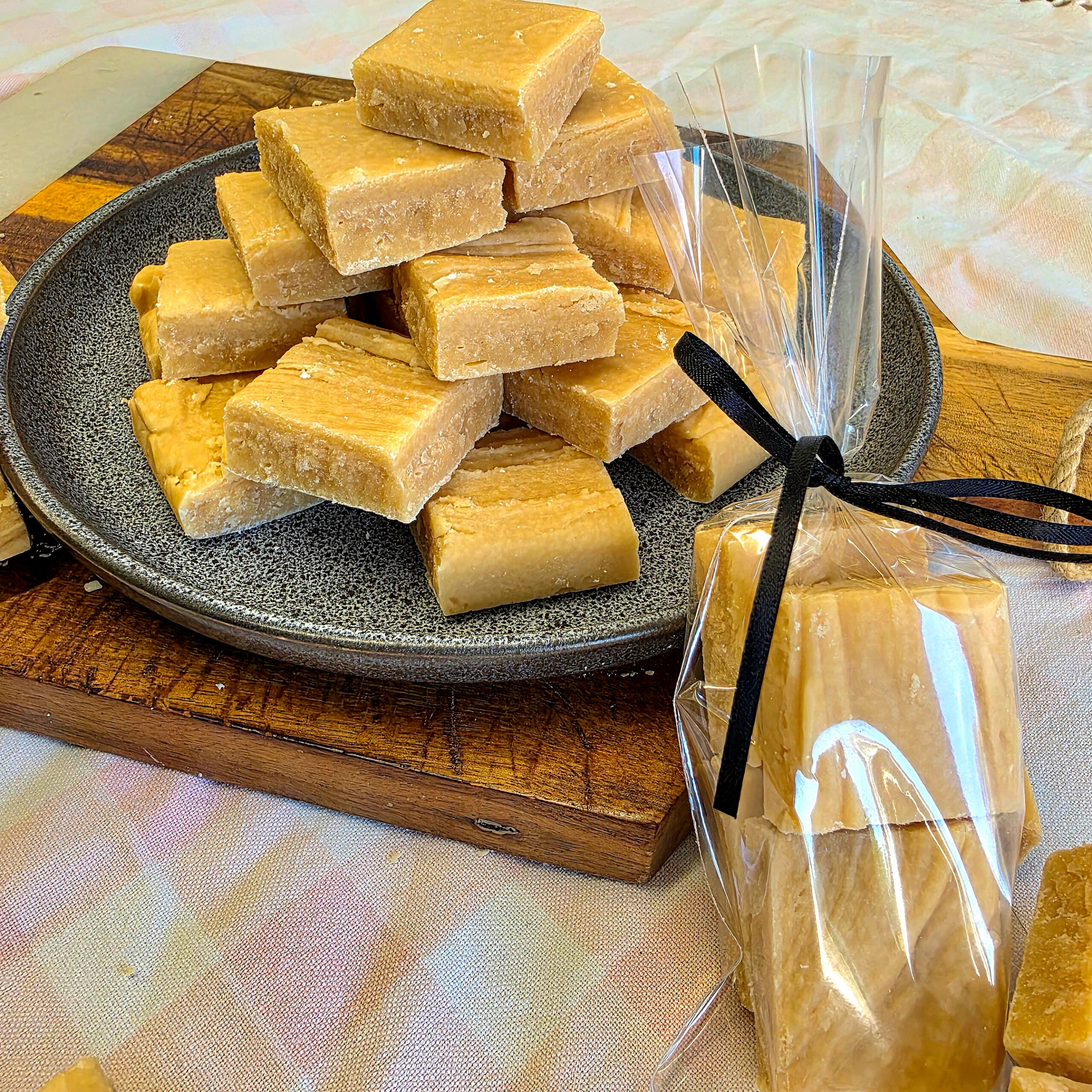 A stack of square pieces of homemade fudge on a plate, with several pieces in a clear gift bag tied with a black ribbon, set on a wooden chopping board.