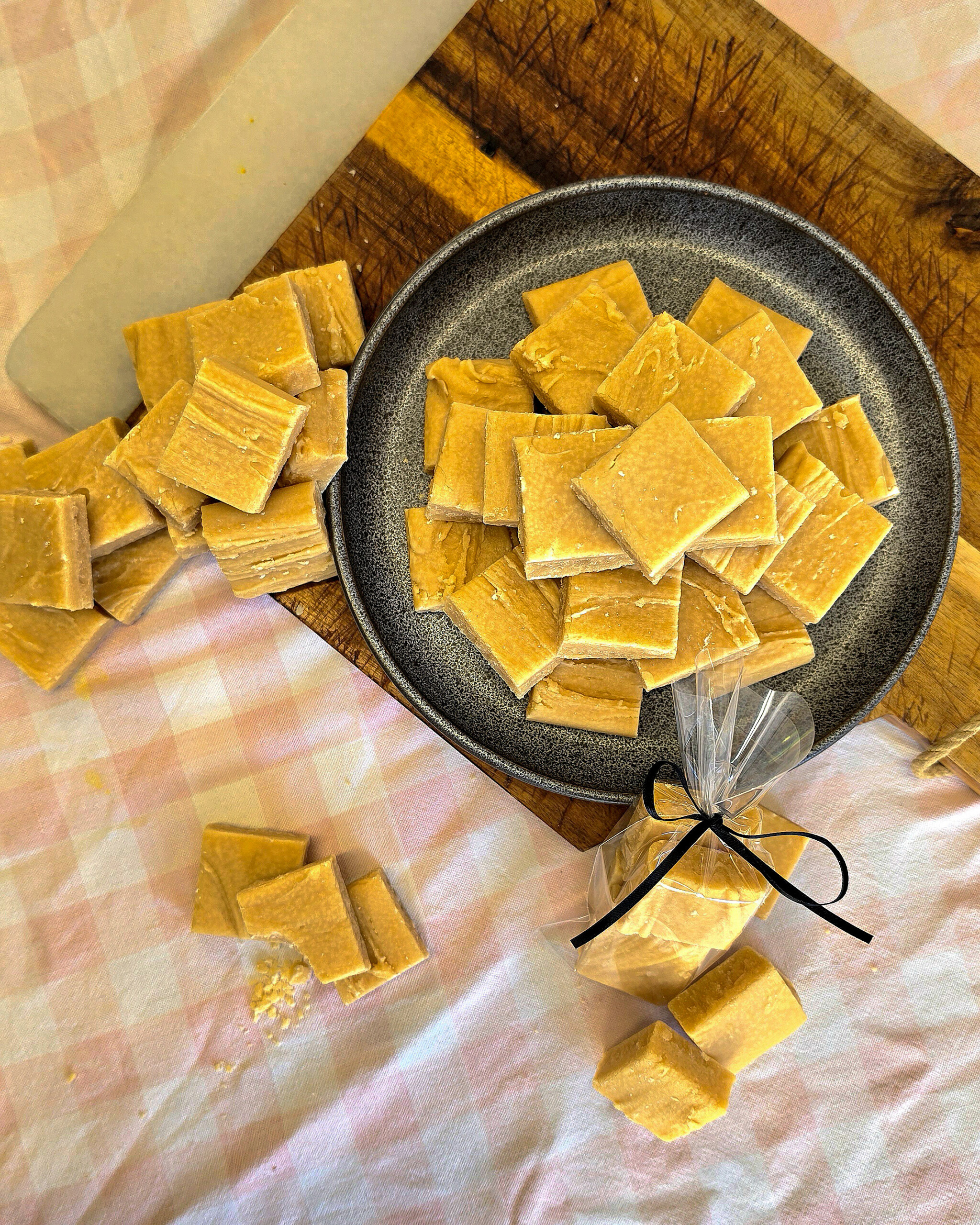 A plate and chopping board with square pieces of reminiscent of classic Scottish tablet, some scattered on a pink checked tablecloth. A small bag of fudge tied with a black ribbon is also shown.