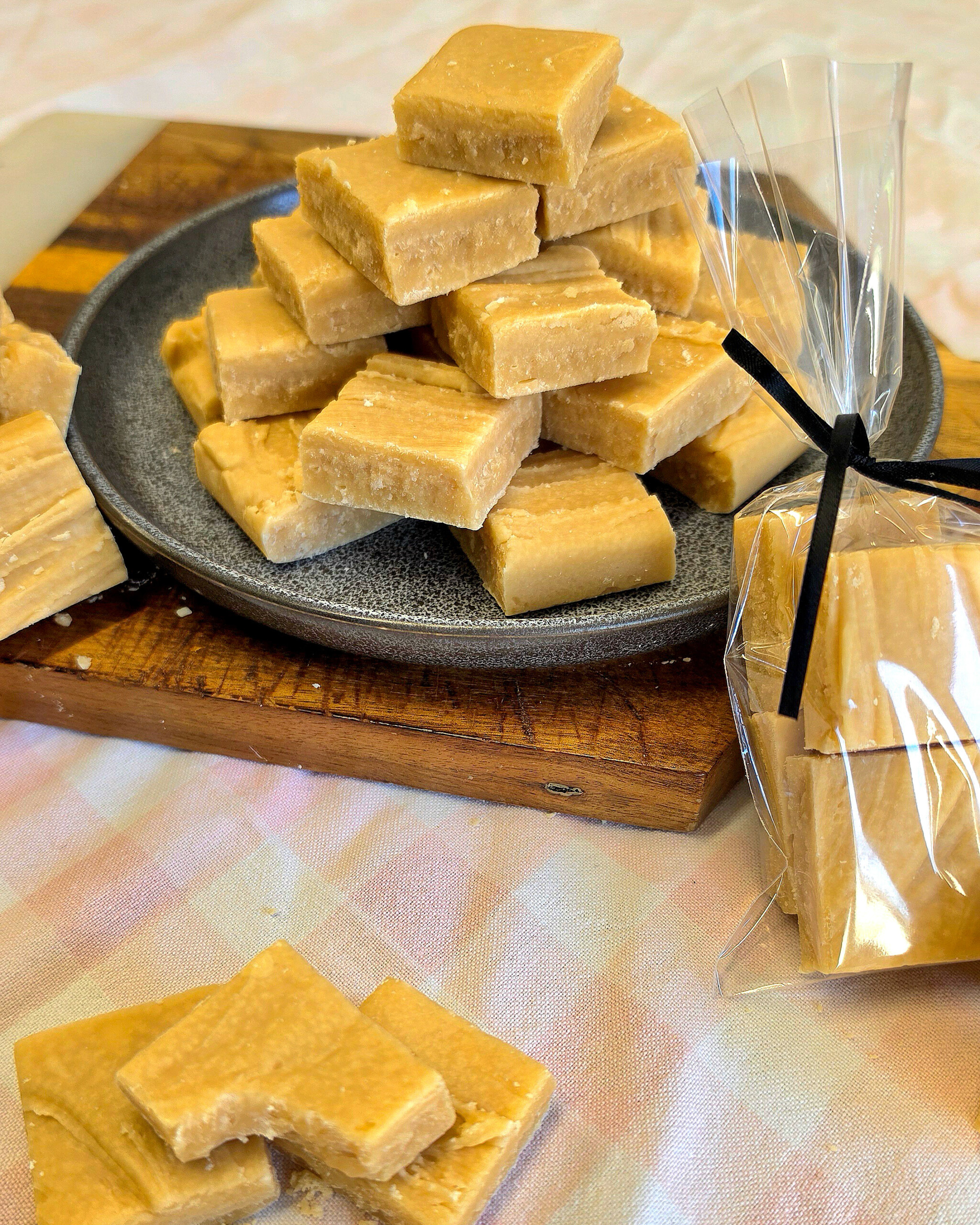 Squares of homemade reminiscent of classic Scottish tablet are stacked on a grey plate, with more pieces nearby and some packaged in a clear plastic bag tied with a black ribbon, all set on a wooden board and a pastel tablecloth.