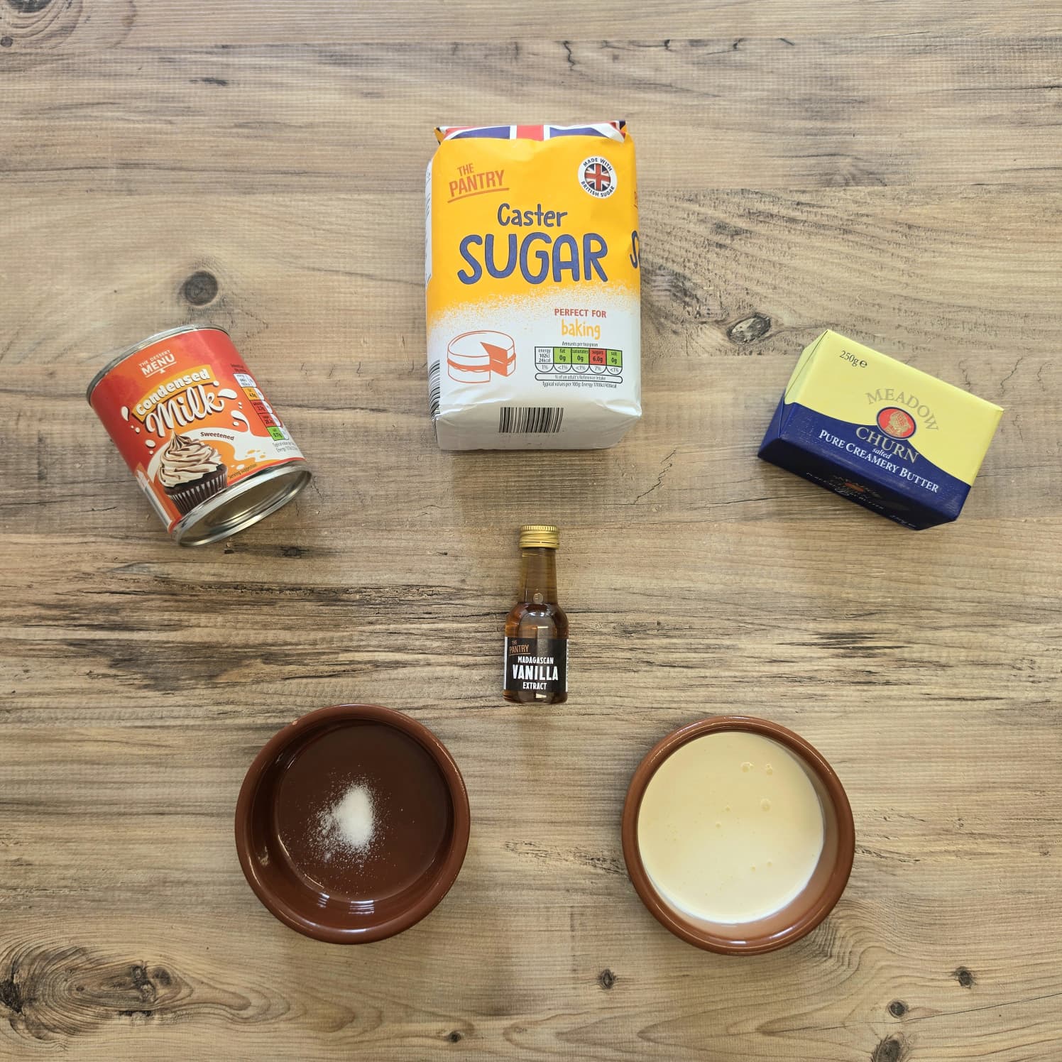 Five baking ingredients on a wooden table: condensed milk, caster sugar, butter, vanilla essence, and two small bowls containing salt and a pale liquid.