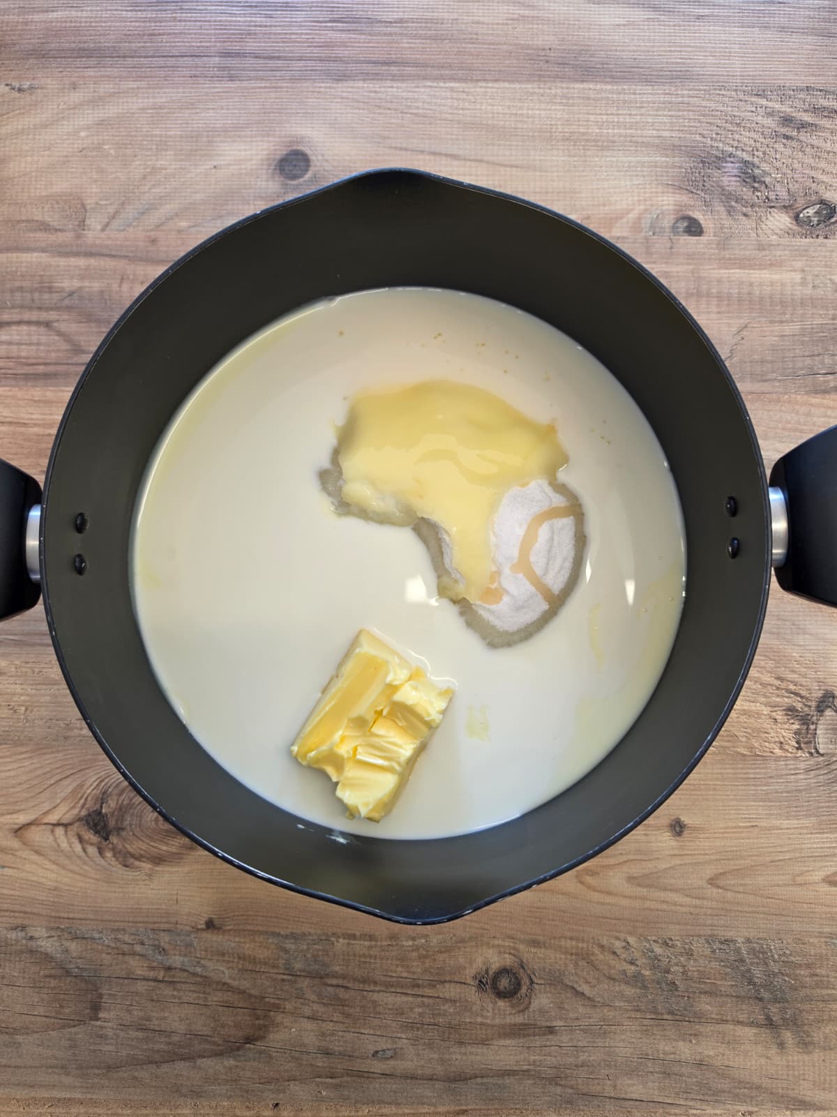 A black saucepan on a wooden surface contains milk, butter, and sugar, all unmixed and ready for cooking.