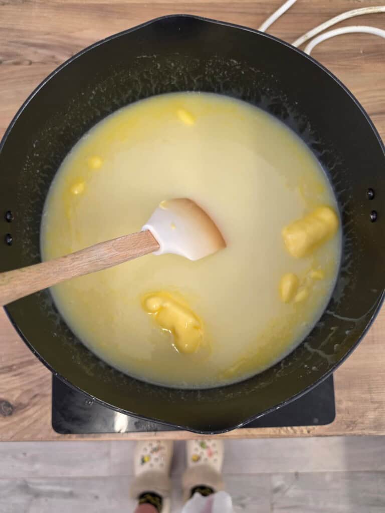 A pot on a hob contains a yellowish liquid with chunks of butter. A spatula with a wooden handle is stirring the mixture. A pair of feet in patterned shoes is visible at the bottom.