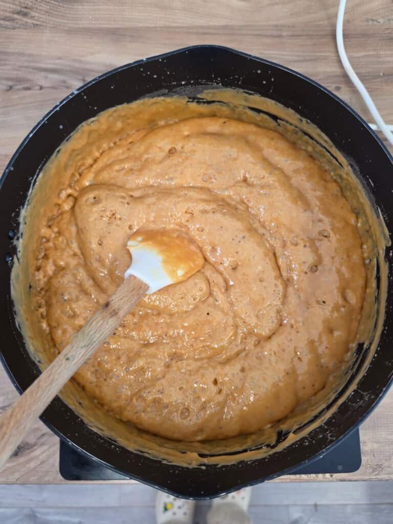 A pot filled with thick, orange batter being mixed with a wooden and silicone spatula on a wooden worktop.