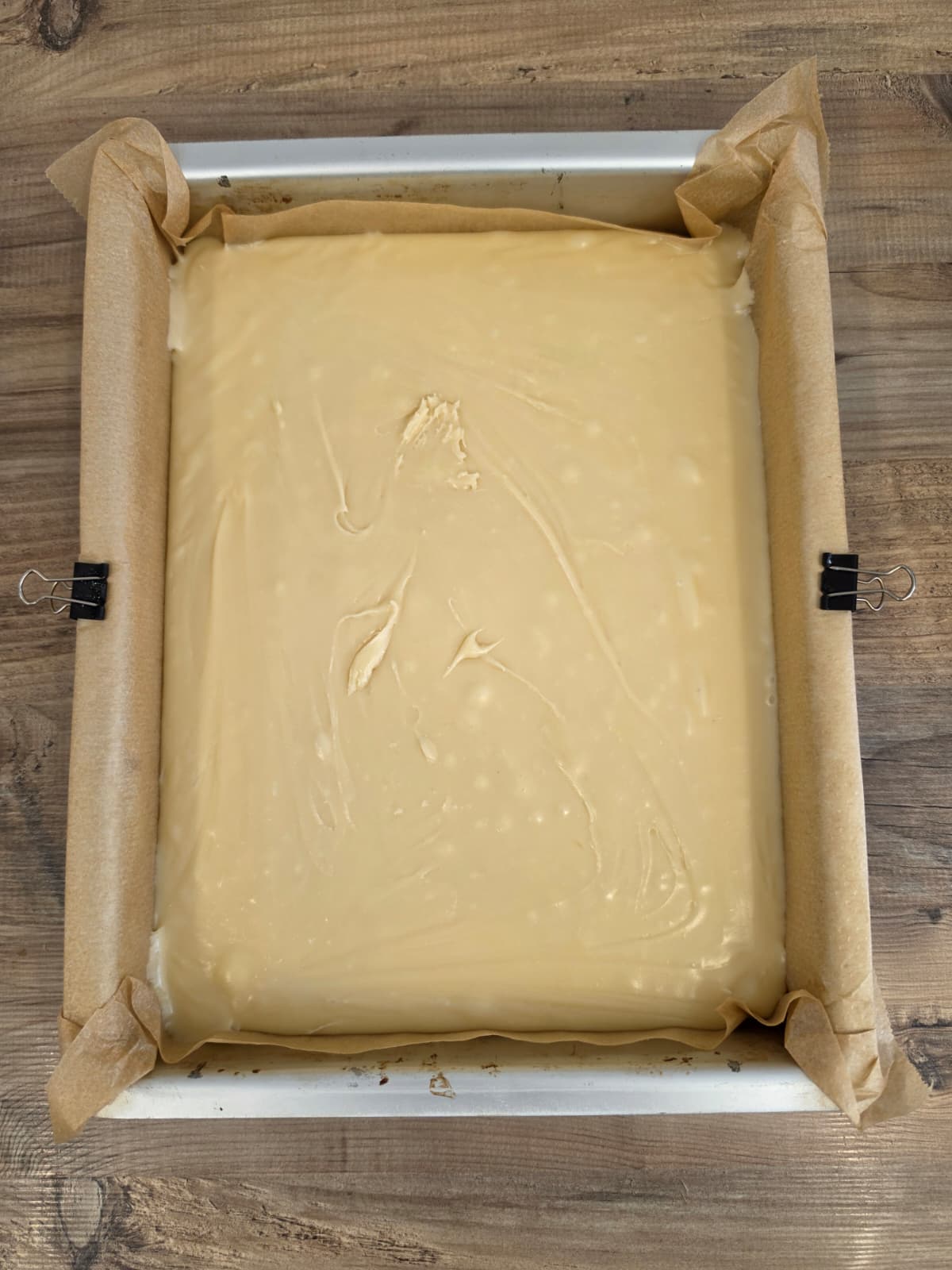 A rectangular baking tin lined with greaseproof paper and filled with smooth, light scottish tablet, placed on a wooden surface. Two bulldog clips secure the greaseproof paper.