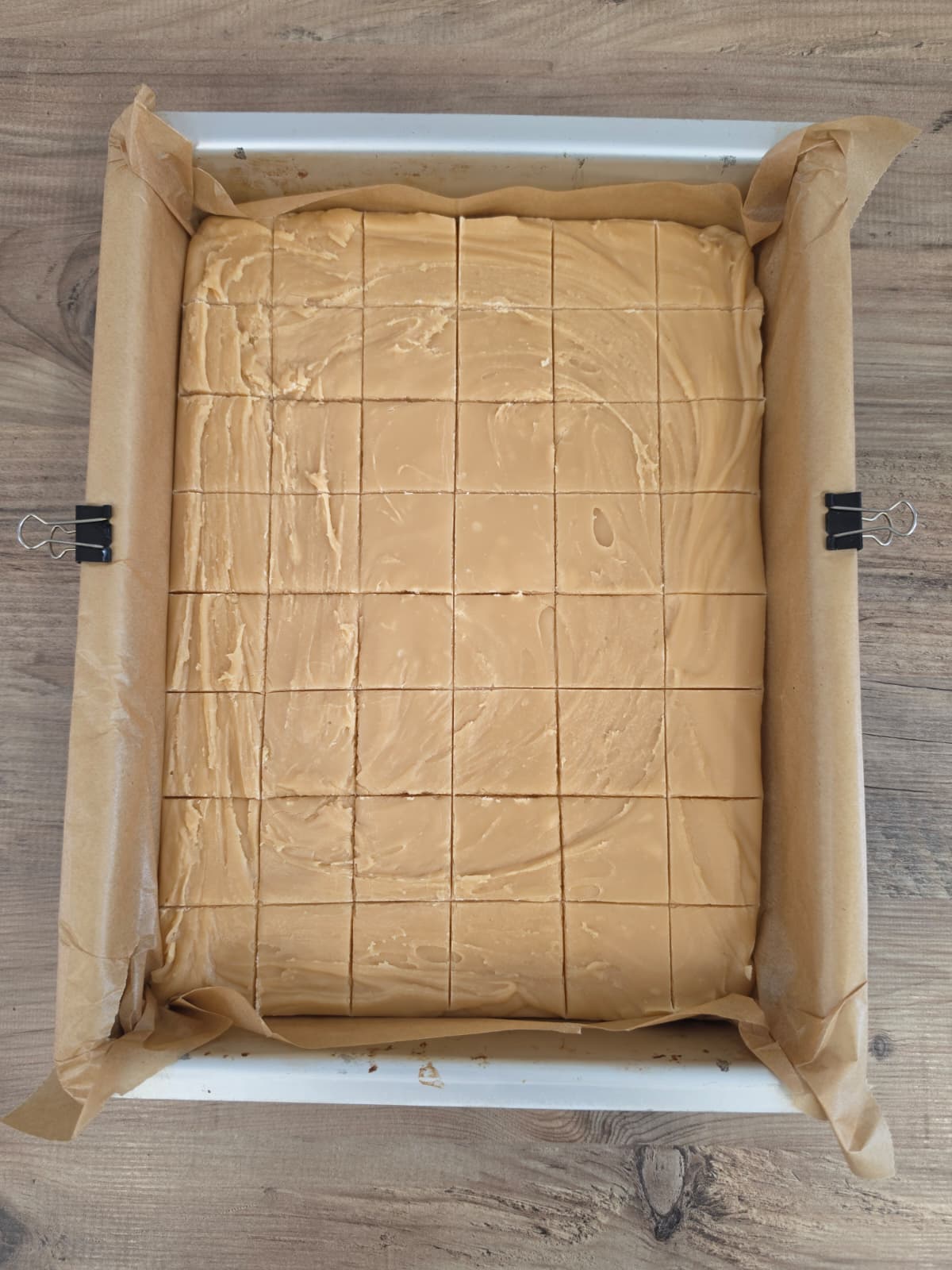 A tray lined with baking parchment holds caramel fudge, cut into neat squares. Two bulldog clips secure the parchment to the tray. The wooden surface below is visible around the tray.