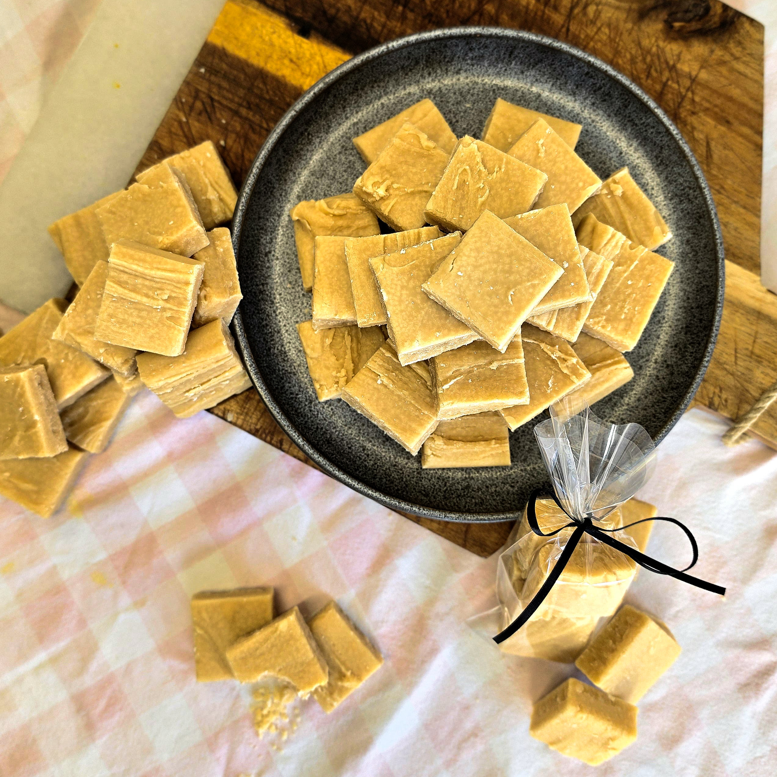 A plate of classic Scottish tablet pieces on a wooden board, with more tablet scattered around and some wrapped in a clear bag tied with a black ribbon.