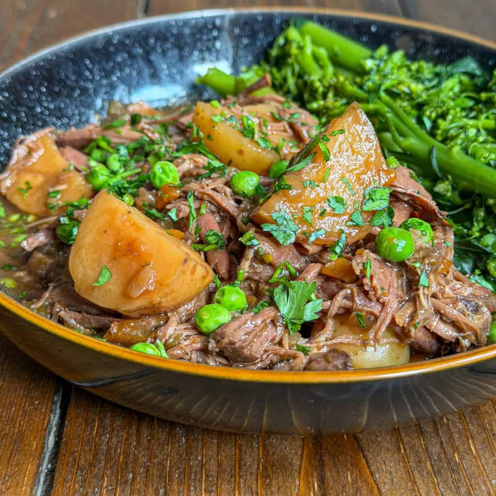 A close-up of a bowl filled with slow cooker beef brisket stew, featuring tender shredded meat, large potato chunks, and green peas, garnished with fresh herbs. A side of bright green broccolini sits in the bowl on a rustic wooden table.