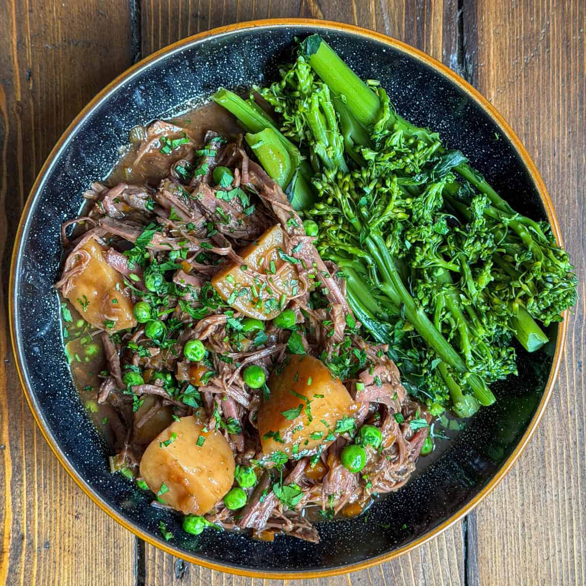 A bowl of slow cooker beef brisket braised with potatoes, garden peas, and herbs in a rich brown sauce, served with a side of steamed tenderstem broccoli on a rustic wooden table.