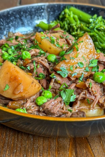 A close-up of a bowl filled with slow cooker beef brisket stew, featuring tender shredded meat, large potato chunks, and green peas, garnished with fresh herbs. A side of bright green broccolini sits in the bowl on a rustic wooden table.