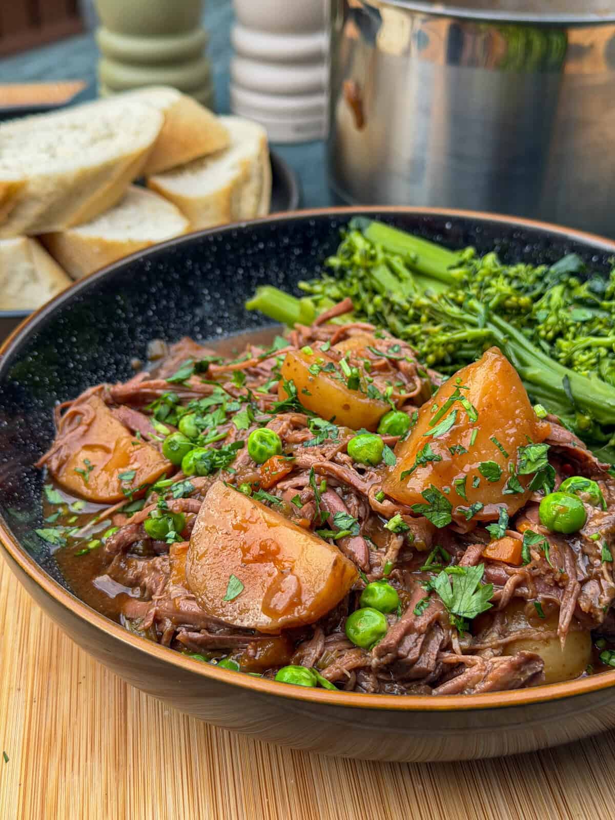 A bowl of slow cooker beef brisket stew with potatoes, peas, and herbs, served with steamed tenderstem broccoli. Slices of bread are visible in the background. The food is arranged on a wooden surface.