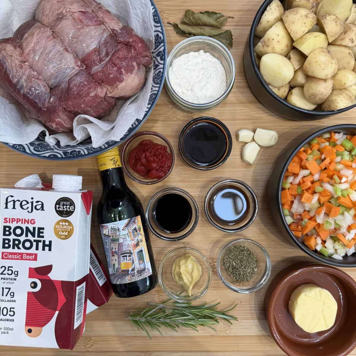 Ingredients for a hearty slow cooker beef brisket stew are arranged on a wooden surface, including raw beef, bone broth, potatoes, herbs, vegetables, tomato purée, mustard, soy sauce, balsamic vinegar, flour, garlic and bay leaves.