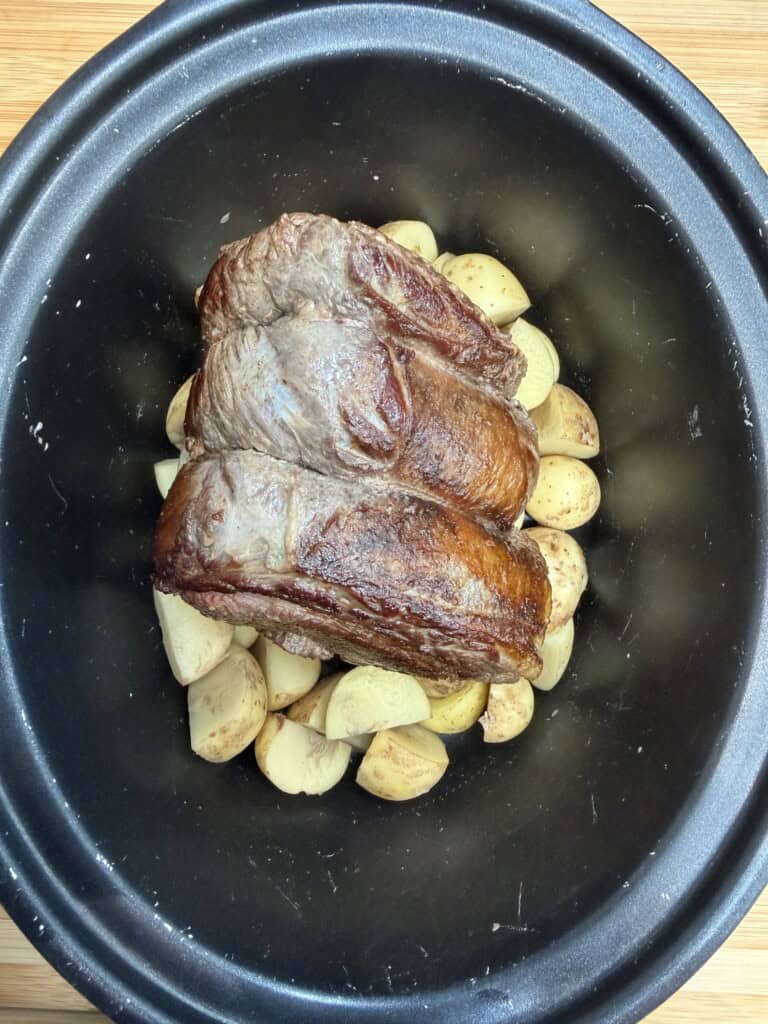 A browned beef roast sits atop quartered potatoes in a black slow cooker, ready to be cooked.