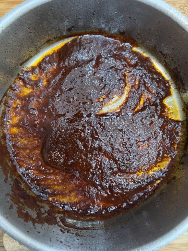 A top-down view of a round metal saucepan containing a thick, dark brown, caramelised sauce that covers the bottom, leaving some yellowish, oily residue around the inner edges of the saucepan.