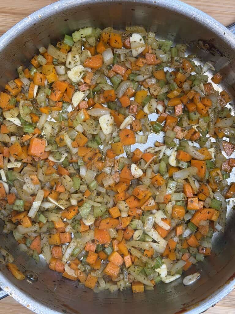 Chopped carrots, onions, celery, and garlic sautéing in a large metal pan, seasoned with herbs and spices. The vegetables are lightly browned and mixed evenly.