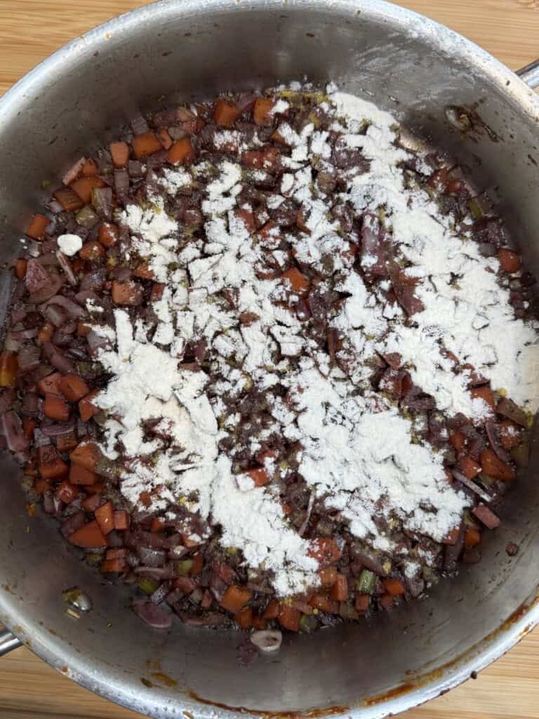 Diced carrots, onions, and celery are sautéing in a pan, topped with a layer of white flour, likely in preparation for a stew or sauce.