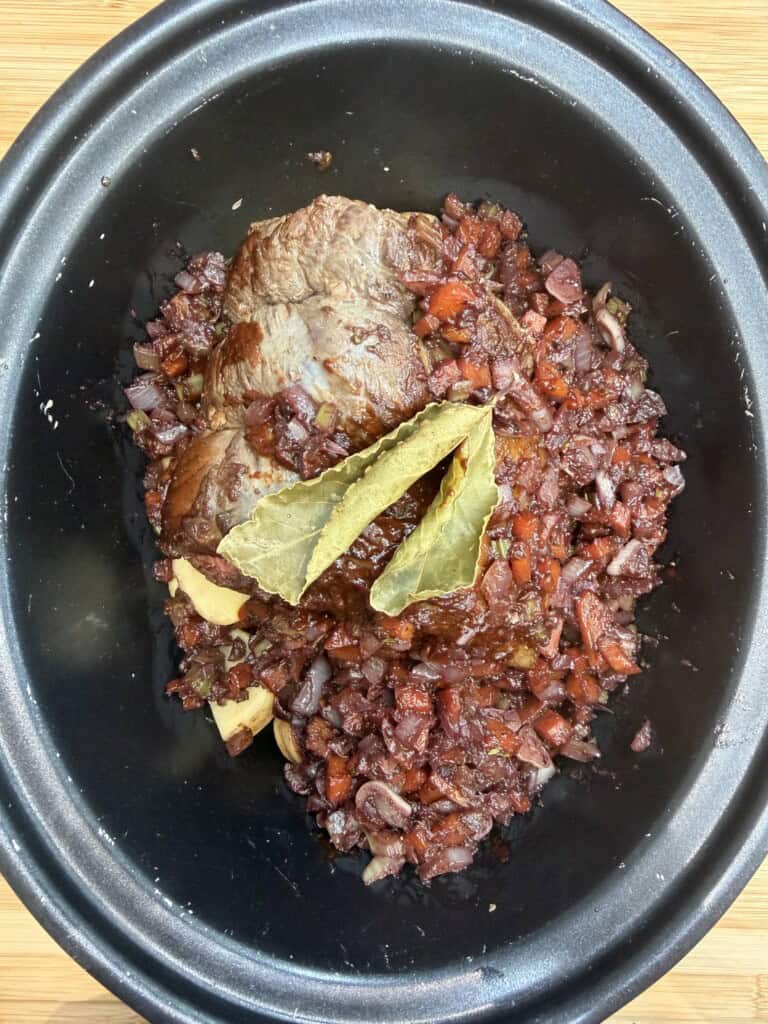 Top-down view of a slow cooker bowl containing a browned beef joint, diced vegetables in a dark sauce, and two bay leaves resting on top. Beneath the joint, pieces of potato are visible. The pot sits on a wooden surface.