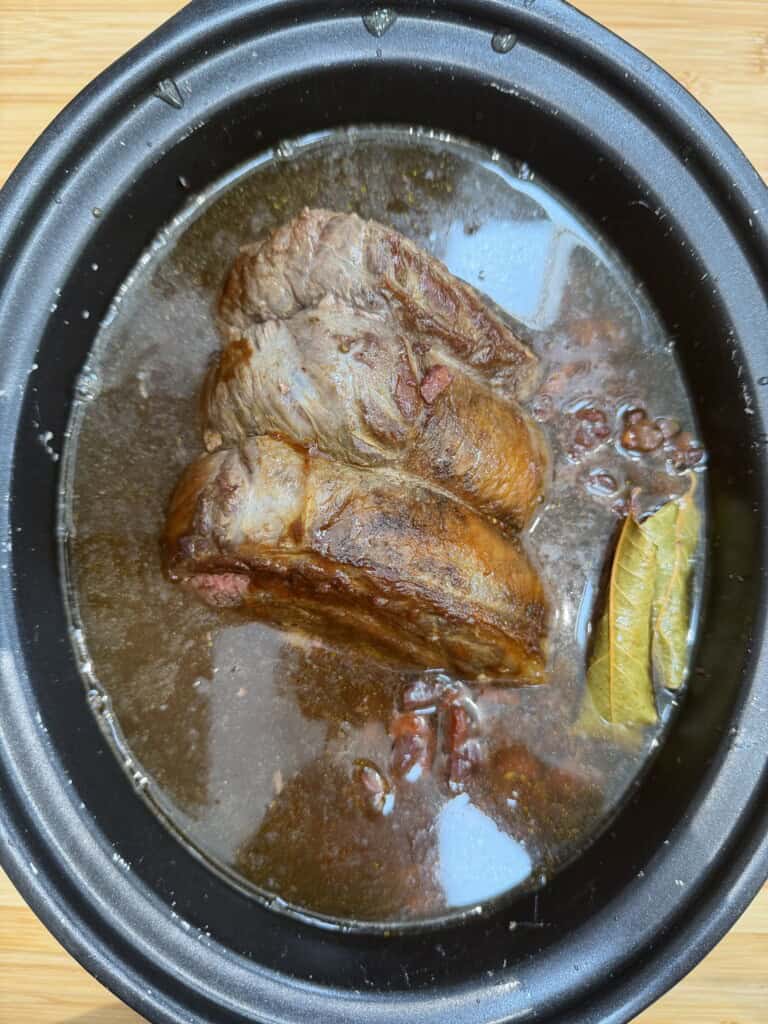 A large piece of beef brisket is simmering in dark stock with beans and a bay leaf inside a black slow cooker.