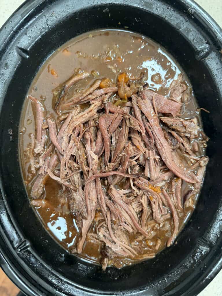 A top-down view of tender, shredded slow cooker beef brisket cooking in a dark, rich broth inside a black slow cooker, with small pieces of carrots and onions visible in the liquid.