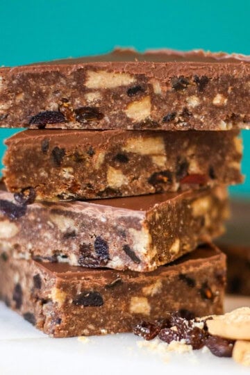 A stack of four rectangular chocolate tiffin biscuit bars topped with a smooth layer of chocolate, with loose sultanas and biscuit pieces on the surface below.