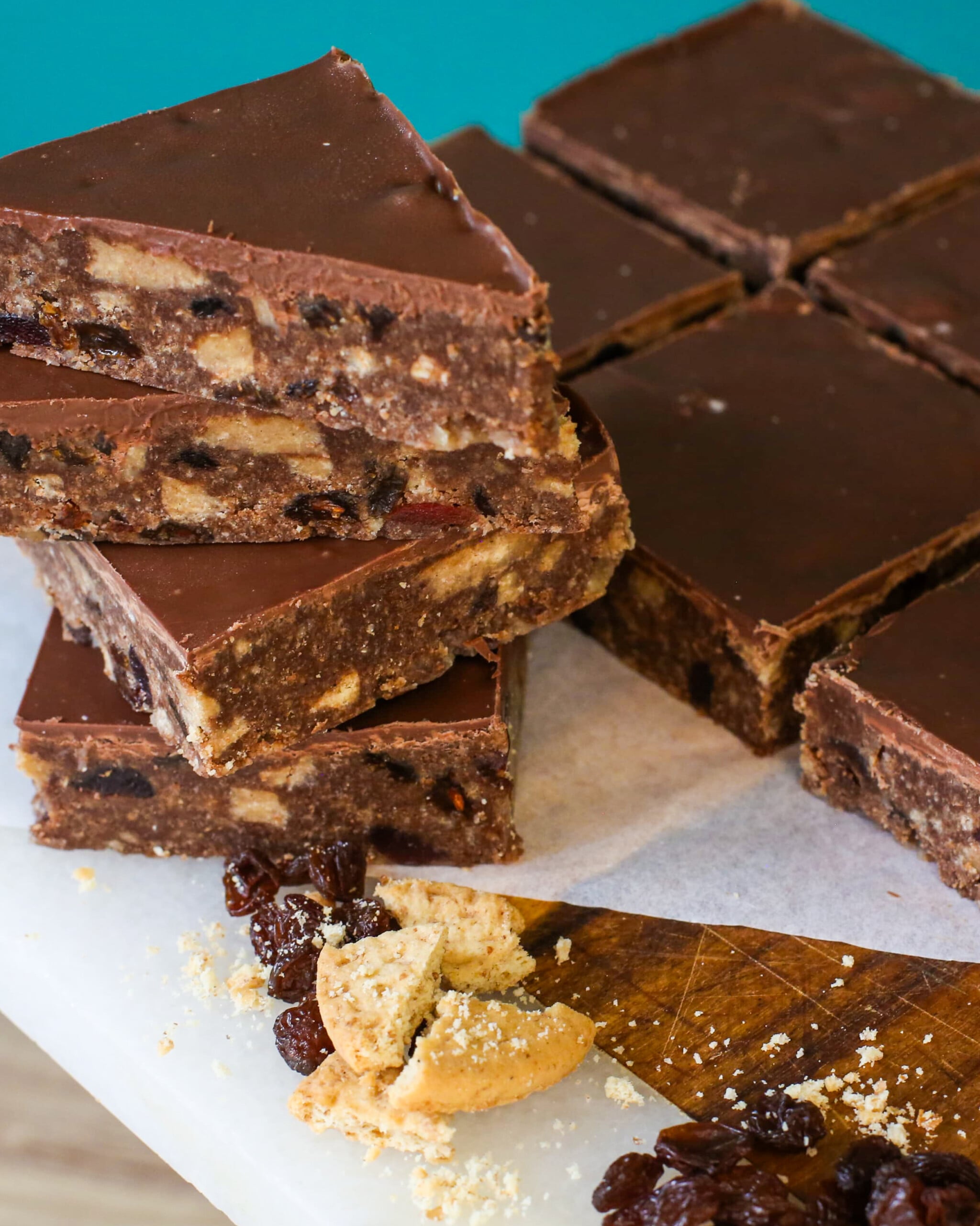 A stack of chocolate tiffin bars topped with a smooth ganache, next to more bars, loose sultanas, and broken biscuit pieces on a white surface.