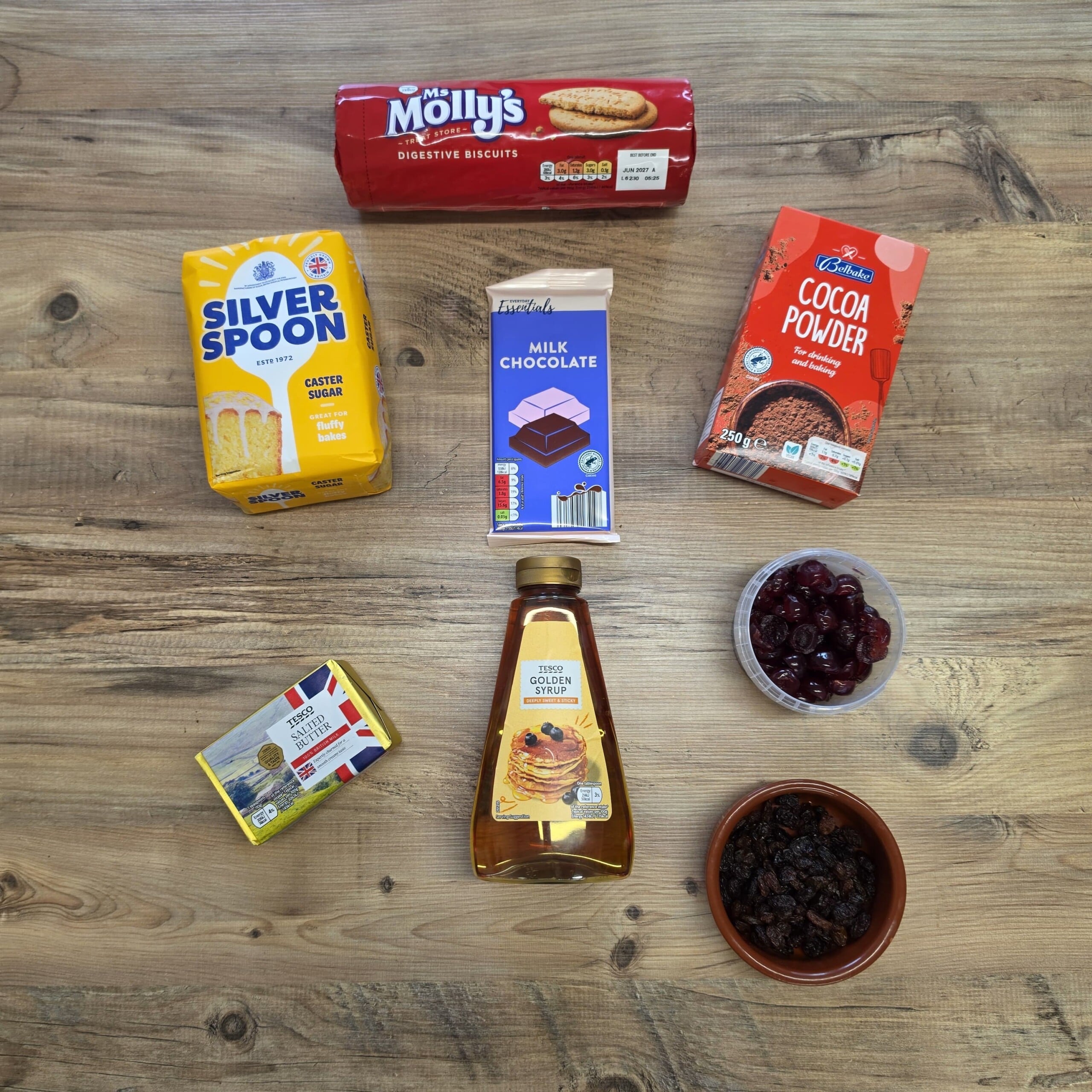 A flat lay of baking ingredients on a wooden surface, including a packet of digestive biscuits, caster sugar, milk chocolate, cocoa powder, salted butter, golden syrup, dried cherries, and sultanas.
