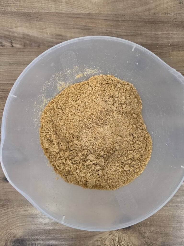 A pile of brown, sandy crumbs in a clear plastic mixing bowl on a wooden surface.