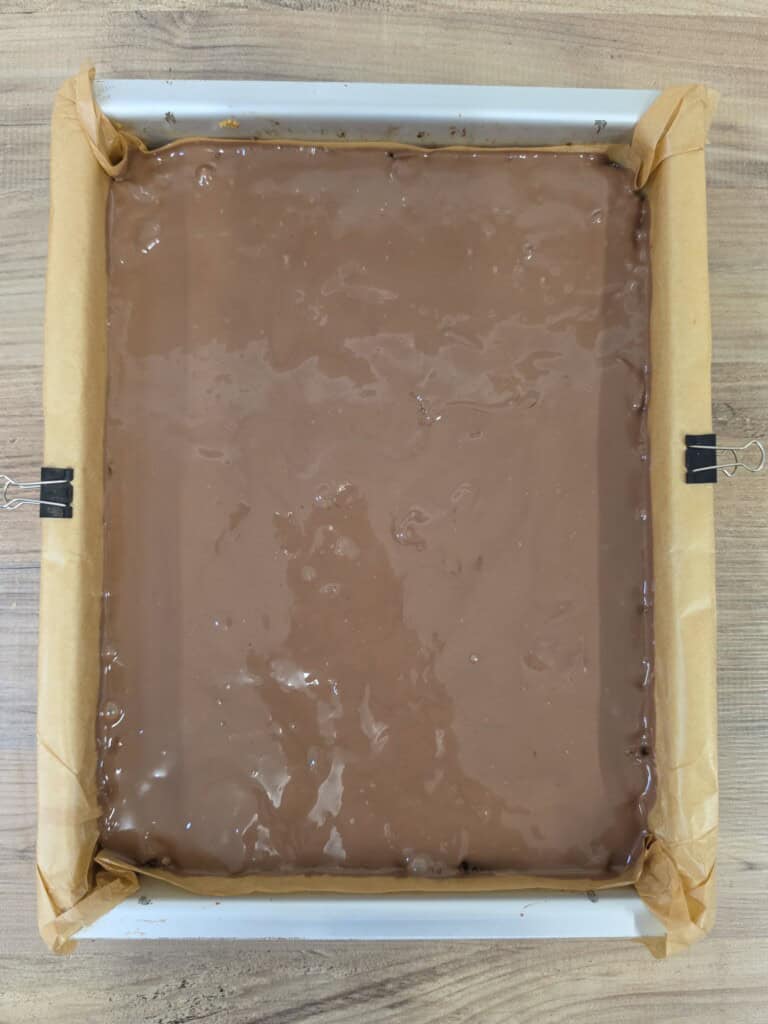 A metal baking tin lined with greaseproof paper containing a smooth, poured layer of milk chocolate.
