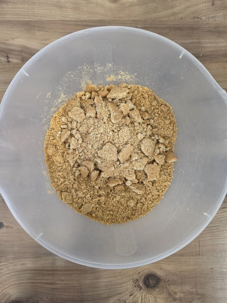 A clear plastic bowl containing a mixture of crushed digestive biscuit crumbs and small biscuit chunks.
