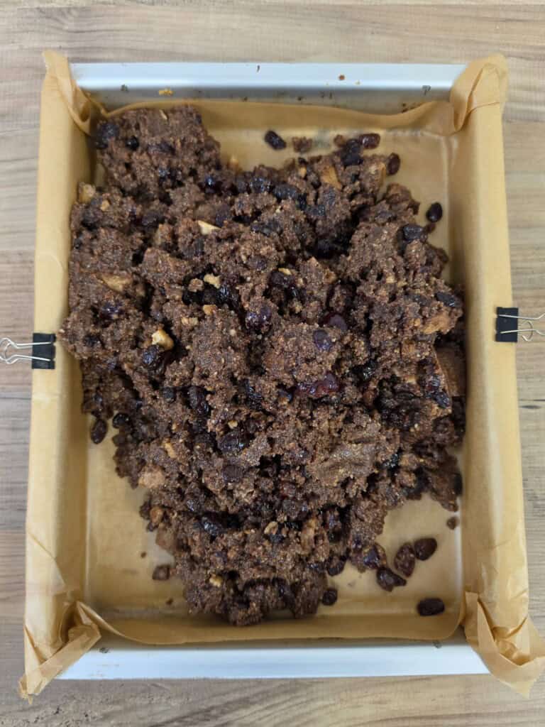 A baking tray lined with greaseproof paper containing a mound of dark, crumbly cake mixture mixed with dried fruit.