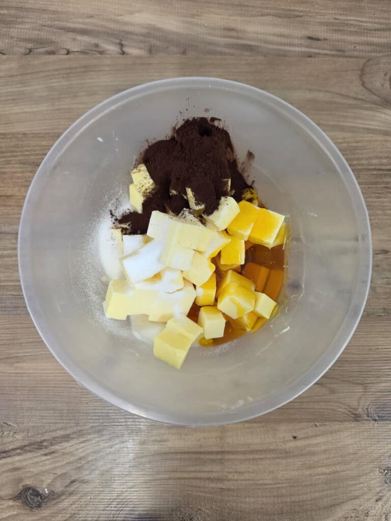 A clear mixing bowl containing cubed butter, white sugar, cocoa powder, and golden syrup on a wooden surface.