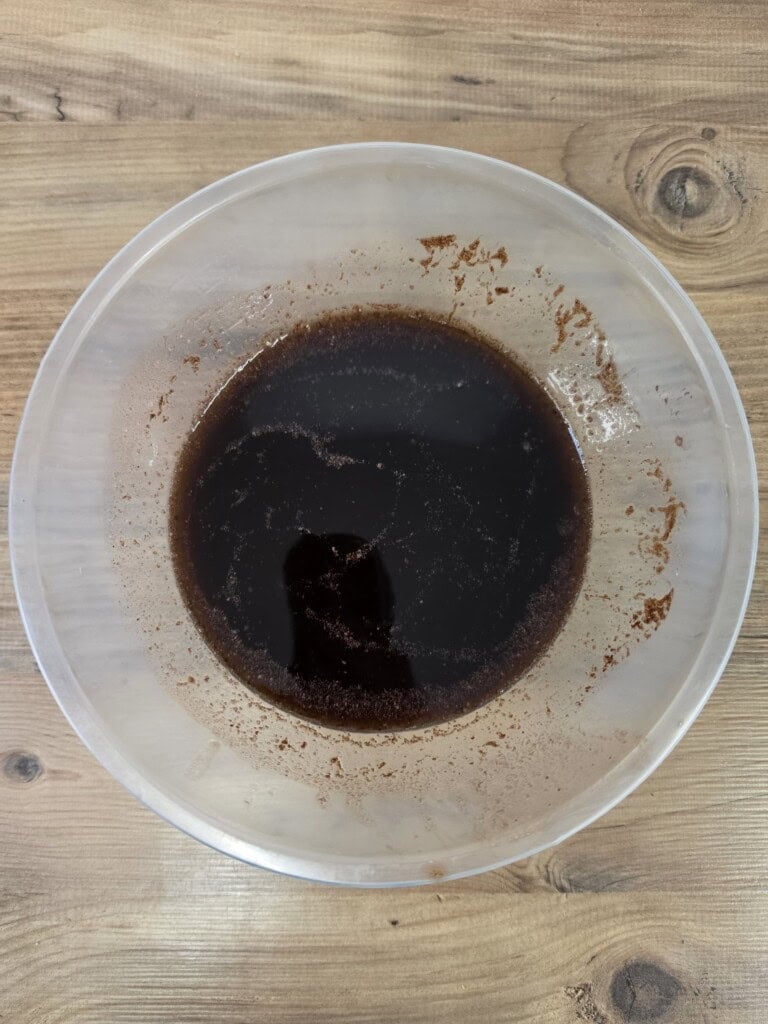 A clear plastic mixing bowl containing dark brown liquid on a wooden surface.