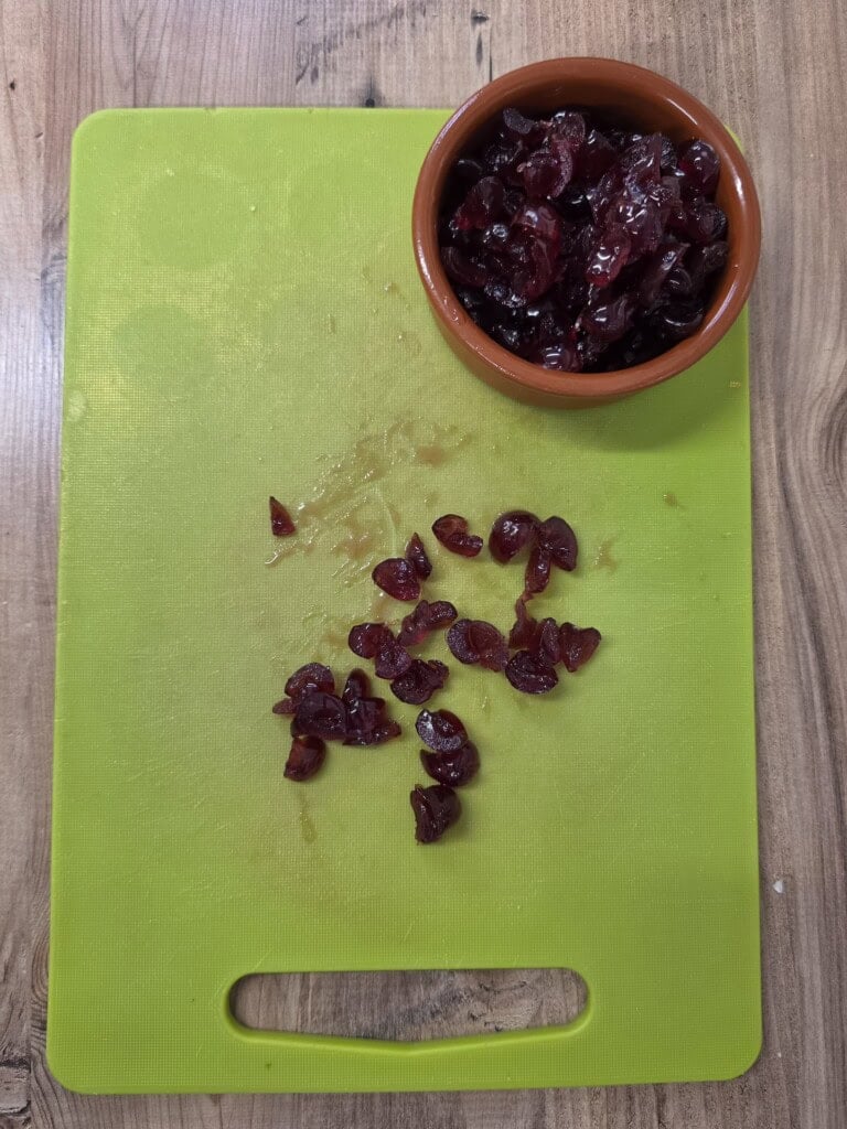 A small bowl of halved cherries sits on a green chopping board, with a few loose cherries scattered below.