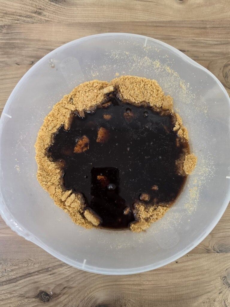 A clear mixing bowl containing a circle of crushed digestive biscuit crumbs surrounding a pool of dark liquid.